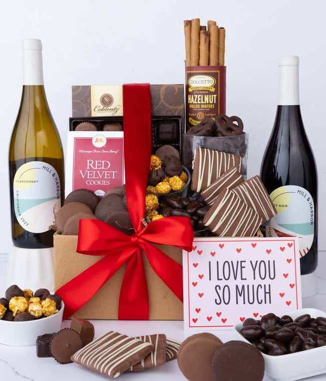 A gift box overflowing with treats including an assortment of chocolate-covered items like oreoes, almonds graham crackers and pretzels along with red velvet cookies, hazelnut wafers, an 8 piece box of chocolates, 2 bottles of wine and an I Love You card.