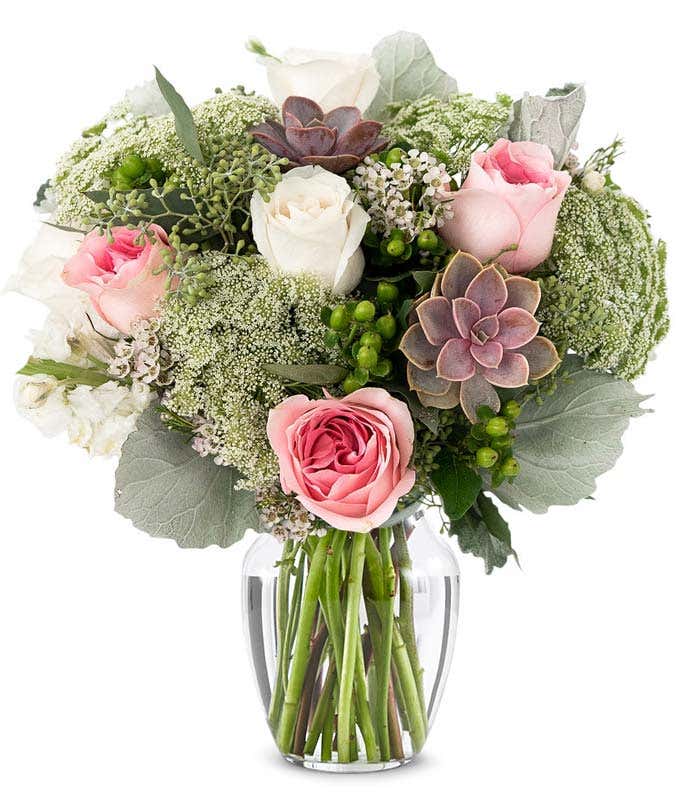 A floral arrangement featuring white roses, pink roses, succulents, white stock, hypericum berries, Queen Anne's lace, white waxflower, and seeded eucalyptus, with an optional glass vase.