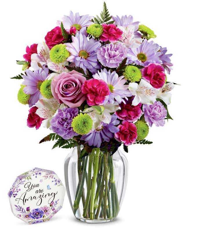 A bouquet featuring lavender daisies, lavender roses, hot pink mini carnations, lavender carnations, white alstroemeria, and green button poms, with an optional vase. Next to a nonagon clear acrylic keepsake reading 'you're amazing' with purple flowers.