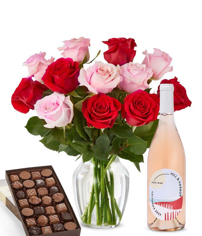 A bouquet of 12 red and pink roses arranged in a glass vase is displayed beside a bottle of Hill & Harbor Rosé and a 1 pound box of assorted chocolates.