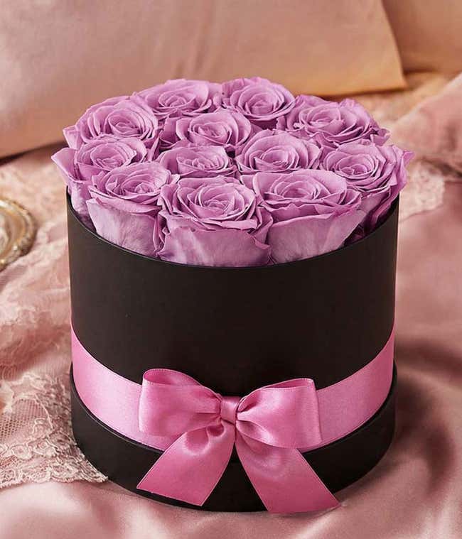 One dozen light purple preserved roses arranged into a black hat box with a purple ribbon.