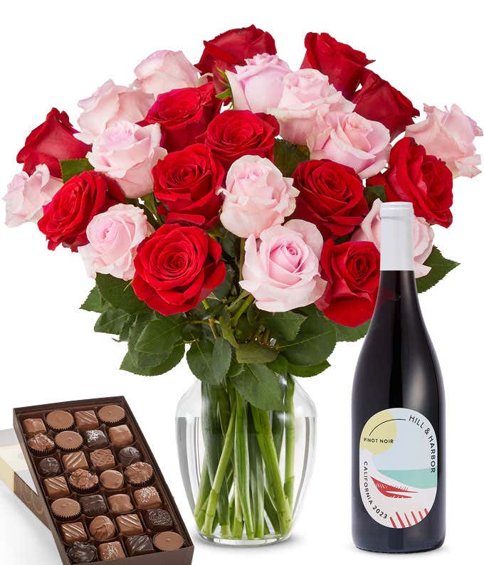 A bouquet of 24 red and pink roses arranged in a glass vase is displayed beside a bottle of Hill & Harbor Pinot Noir and a 1 pound box of assorted chocolates.