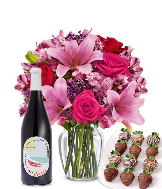 A mixed bouquet of pink and purple flowers in a clear glass vase is displayed with a bottle Hill & Harbor Pinot Noir and 12 chocolate-covered strawberries.