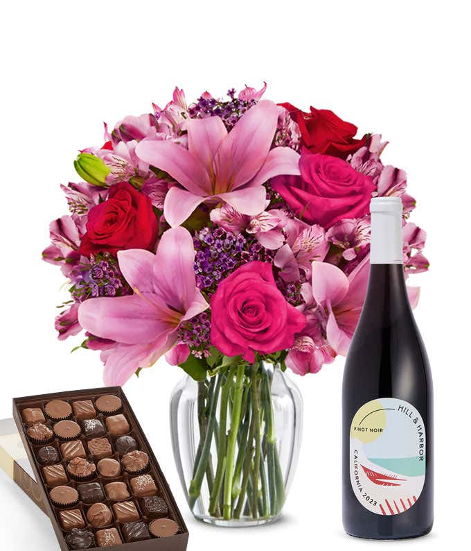 A mixed bouquet of pink and purple flowers in a clear glass vase is paired with a bottle of Hill & Harbor Pinot Noir and a box of assorted chocolates.