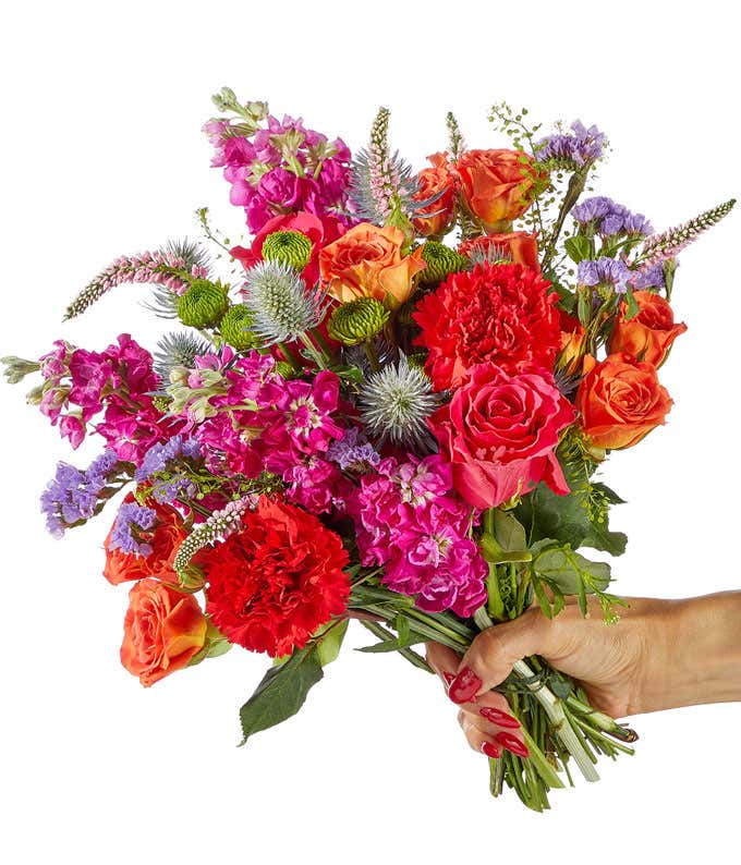A hand-tied bouquet of hot pink roses, red blooms, purple flowers, thistle accents, green filler, and leafy greenery in a fresh and colorful garden-inspired style.