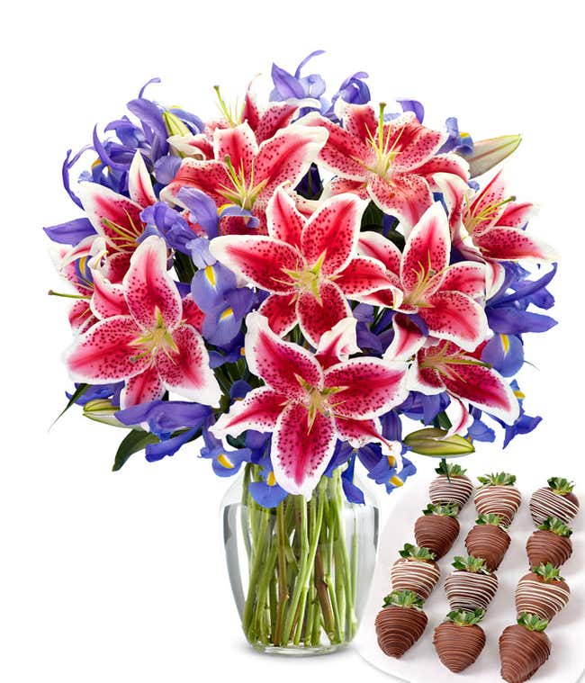 Bouquet of vibrant stargazer lilies and deep purple irises in a clear glass vase, paired with a plate of swizzled chocolate-covered strawberries drizzled in dark and white chocolate.