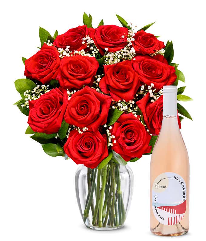 A bouquet of vibrant red roses arranged in a clear glass vase, surrounded by small white flowers and lush green leaves, paired with a bottle of rose wine from California, 2023, featuring a stylish, minimalist label.