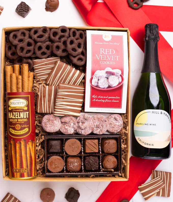 A gift box with chocolate pretzels, chocolate graham crackers, red velvet cookies, hazelnut wafer rolls, an 8 piece box of chocolates and a bottle of sparkling wine.