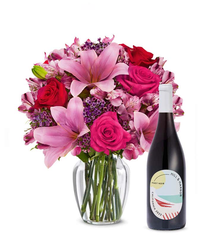 A bouquet of pink lilies, pink roses, and greenery arranged in a clear glass vase sitting next to a bottle of Hill & Harbor California Pinot Noir.