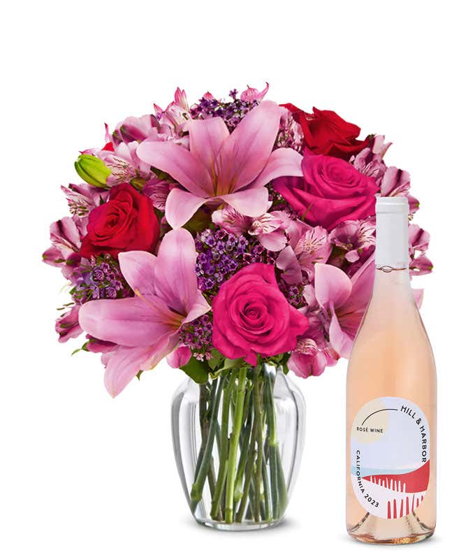 A bouquet of pink lilies, pink roses, and greenery arranged in a clear glass vase sitting next to a bottle of Hill & Harbor California Rosé.