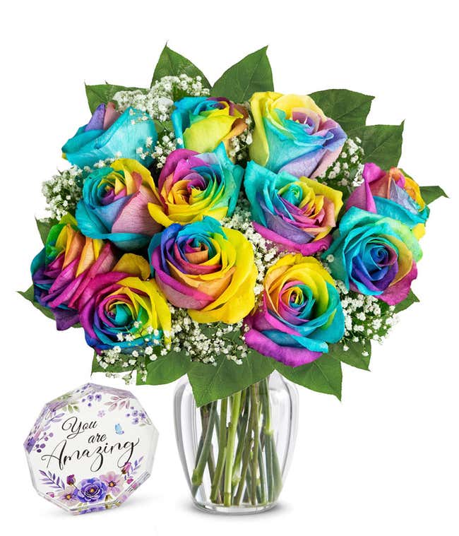 A colorful bouquet of rainbow roses in a clear glass vase, with petals in vibrant hues of blue, yellow, pink, and purple, accented by baby's breath and green leaves, creating a bright and cheerful display.