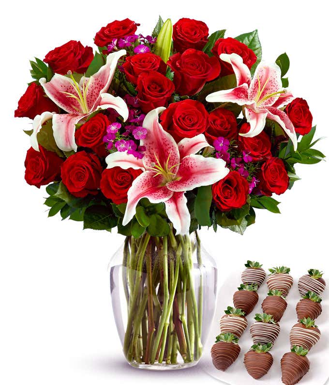 Romantic bouquet of red roses, stargazer lilies, and purple filler flowers in a clear glass vase, paired with chocolate-covered strawberries drizzled in milk and white chocolate.