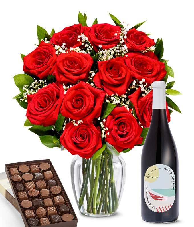 A lush arrangement of a dozen red roses in a clear vase sits beside a bottle of Hill & Harbor Pinot Noir and a 1-pound box of gourmet assorted chocolates.