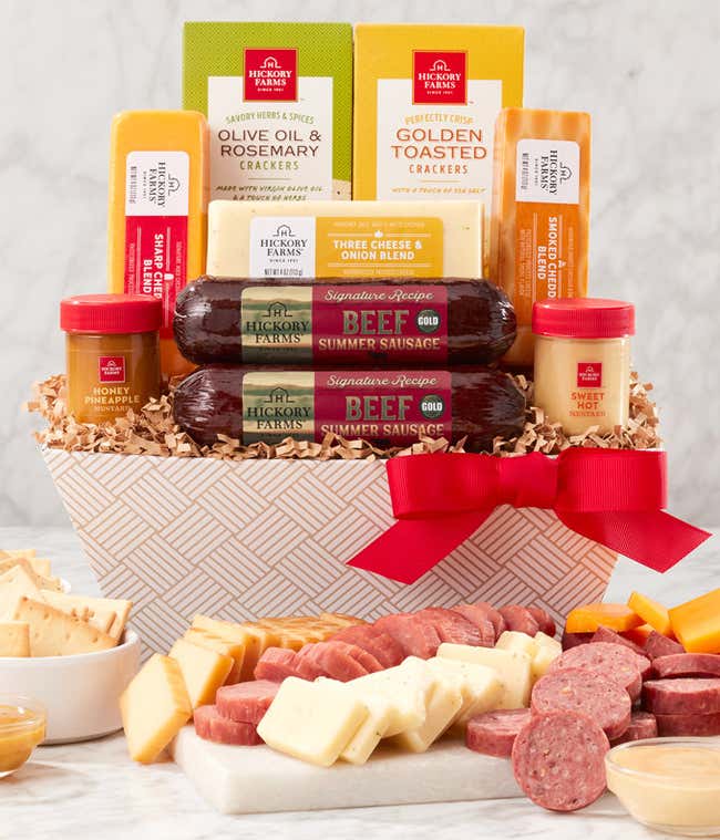 Gift basket with summer sausages, cheeses, crackers, and mustards in a white patterned container with a red bow. A serving board in front displays sliced sausage, cheese, and mustard for dipping.