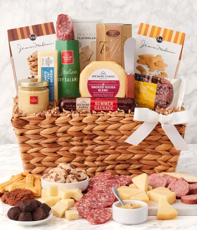 Woven gift basket with salami, cheese, mustard, crackers, cookies, and chocolates. In front, a board displays sliced meats, cheese cubes, and mustard for dipping, with cookies and chocolate truffles arranged alongside. Decorated with a white bow.