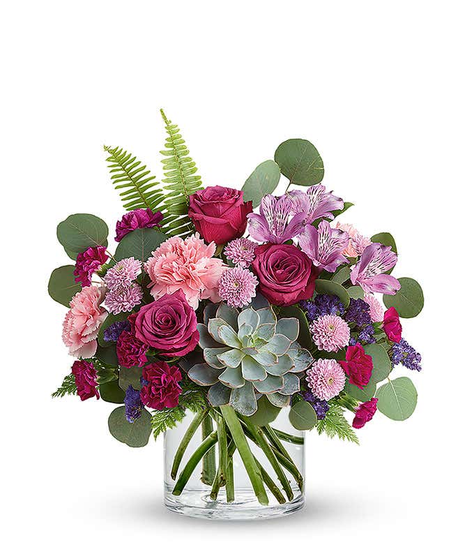Floral arrangement featuring a succulent plant, pink carnations, purple mini carnations, dark pink roses (in deluxe and premium versions), purple statice, purple alstroemeria, lavender button mums, seasonal greenery, and a personal card message in a glass