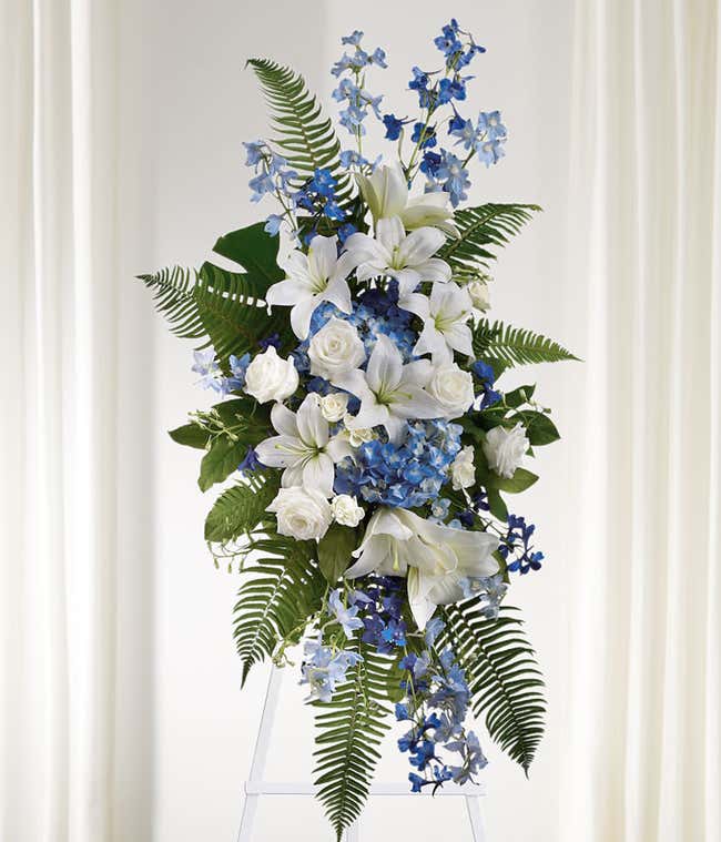 Standing spray with white lilies, roses, blue delphiniums, and green ferns on a white metal easel.