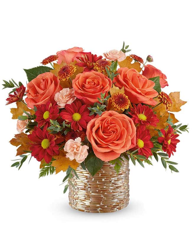 Fall floral arrangement with orange roses, red daisy poms with yellow centers, rust chrysanthemums, light peach carnations, and autumn leaves in a textured gold-glass vase.