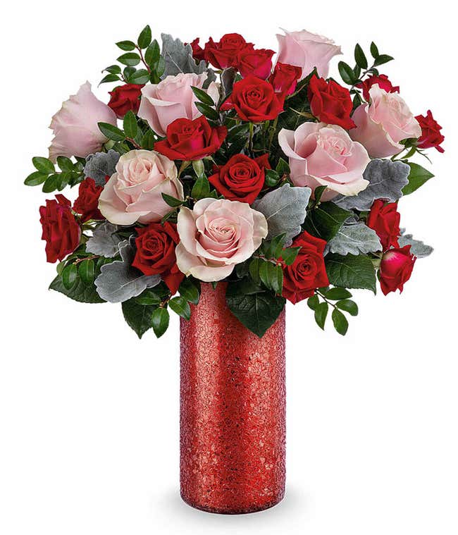 Bouquet of red and blush pink roses with silver dusty miller and lush greenery, arranged in a tall red glitter cylinder vase.