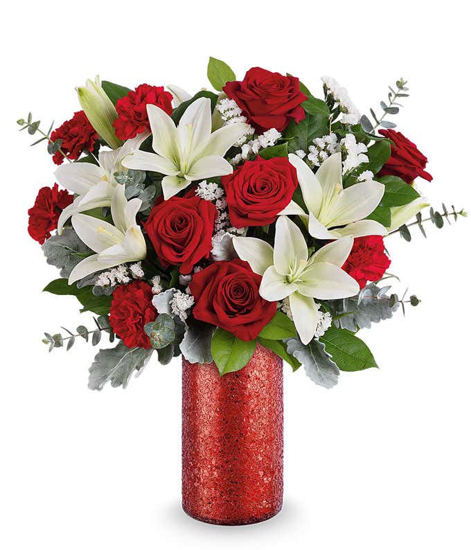 Valentine’s Day arrangement featuring red roses, white lilies, red carnations, silver dusty miller, and lush greenery in a tall red glitter cylinder vase.