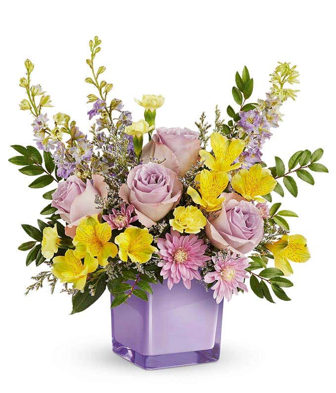 Lavender roses, yellow alstroemeria, mini yellow carnations, lavender larkspur, lavender poms, and floral greenery in a purple cube vase