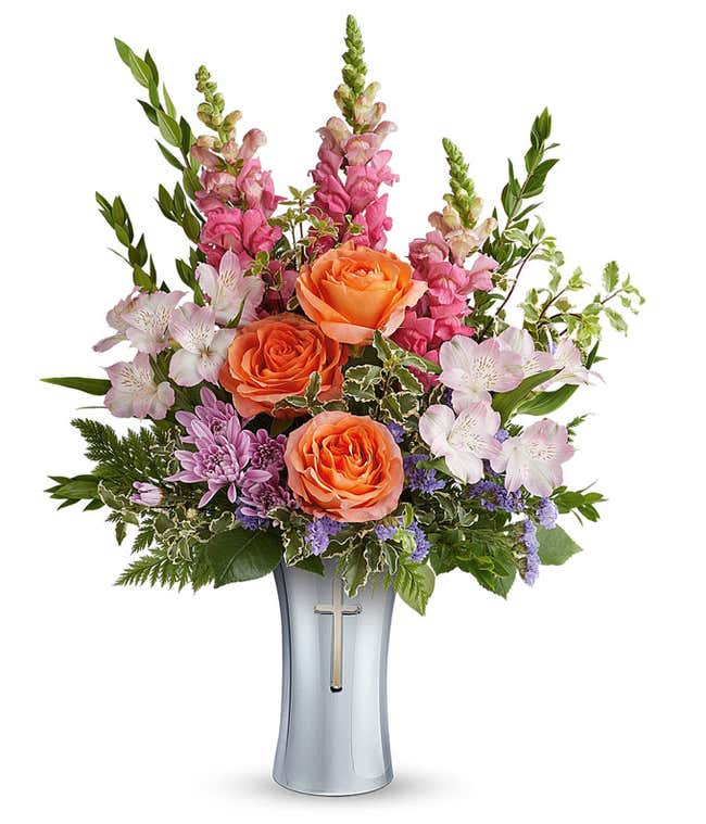 Orange roses, pink snapdragons, pink alstroemeria, lavender cushion spray chrysanthemums, purple statice, and assorted greenery arranged in a keepsake silver cross vase.