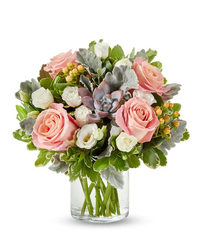 Floral arrangement in a clear glass vase featuring soft pink roses, white spray roses, peach hypericum berries, silver dusty miller, green foliage, and a central mauve-toned succulent.