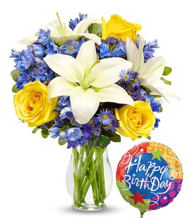 Birthday flower bouquet with white lilies, yellow roses, blue delphinium, and purple asters in a clear glass vase, paired with a colorful Happy Birthday balloon.