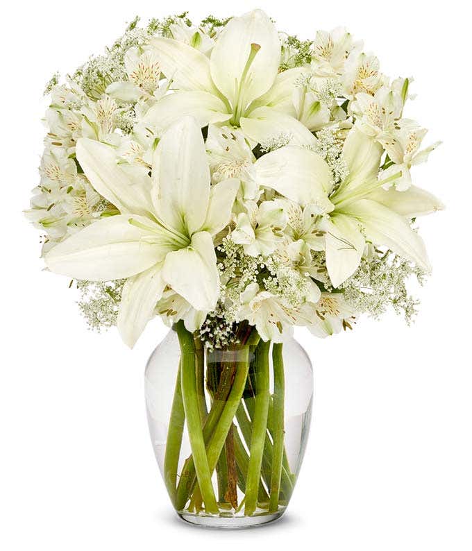 Clear glass vase filled with white lilies, white alstroemeria, and delicate white filler flowers, arranged in a full, rounded bouquet with visible green stems.