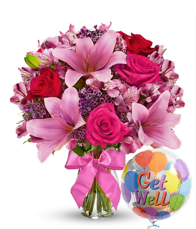 Clear glass vase filled with pink lilies, hot pink roses, red roses, pink alstroemeria, and purple filler flowers, tied with a pink bow and paired with a colorful 'Get Well' balloon. 