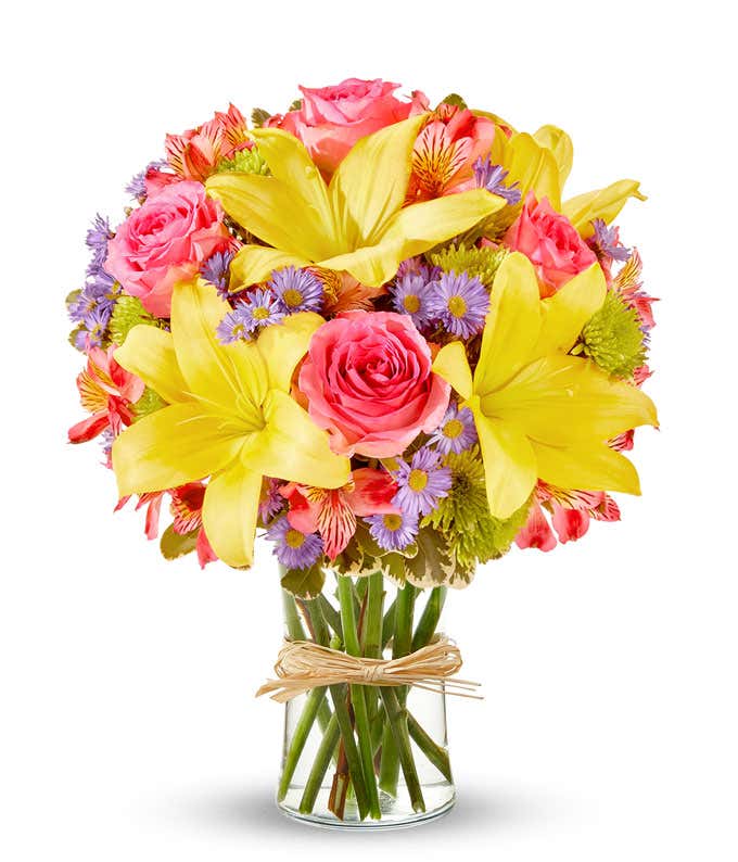 A vibrant arrangement featuring yellow lilies, pink roses, pink alstroemeria, green poms, and purple Monte Casino, beautifully arranged in a glass vase.