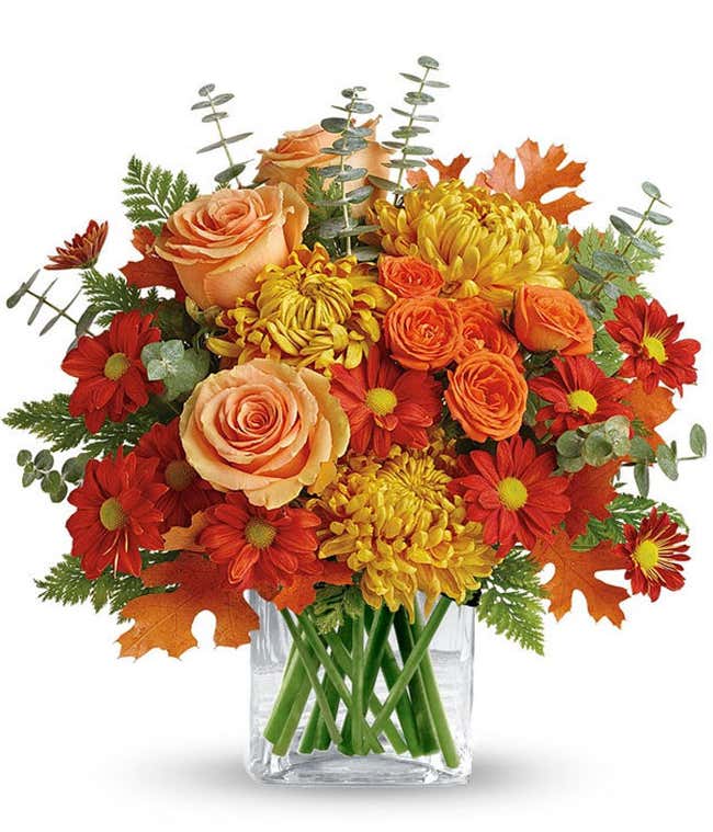 orange roses, yellow chrysanthemums, orange spray roses, red daisy poms, autumn leaves, eucalyptus, and greenery arranged in a clear glass cube vase