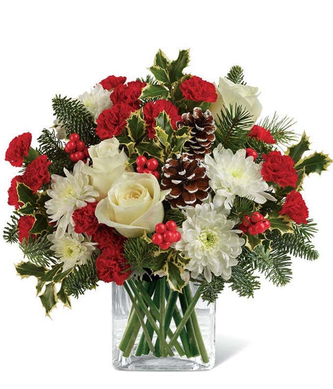 Holiday floral arrangement with white roses, red miniature carnations, white chrysanthemums, pinecones, red berries, holly leaves, evergreen sprigs, and a clear glass cube vase.