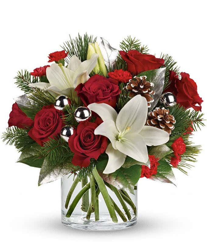 A festive arrangement with red roses, white Asiatic lilies, red mini-carnations, seasonal greens, silver ornaments, and pinecones, beautifully displayed in a clear cylinder vase.