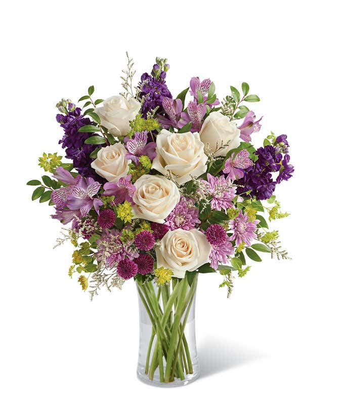 A sophisticated floral arrangement featuring crème roses, lavender alstroemeria, purple stock, purple button chrysanthemums, lavender cushion spray chrysanthemums, white limonium, bupleurum, and huckleberry, all elegantly arranged in a cylinder vase.