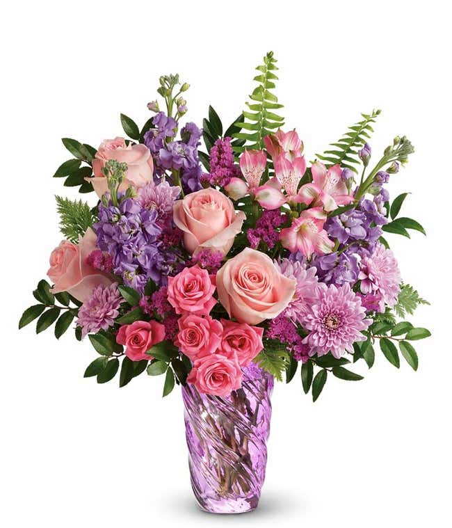 Full bouquet of pink roses, hot pink spray roses, lavender stock, pink alstroemeria, purple poms, and greenery arranged in a lavender swirl glass vase.