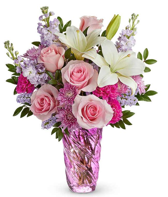 Bouquet of pink roses, white lilies, pink carnations, lavender stock, and purple poms arranged in a twisted lavender glass vase with greenery.