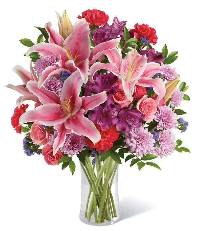 A vibrant bouquet of fresh pink lilies, red carnations, lavender chrysanthemums, purple alstroemeria, pink roses, and greenery arranged in a clear glass vase. The lush mix of colors creates a cheerful and elegant floral display.