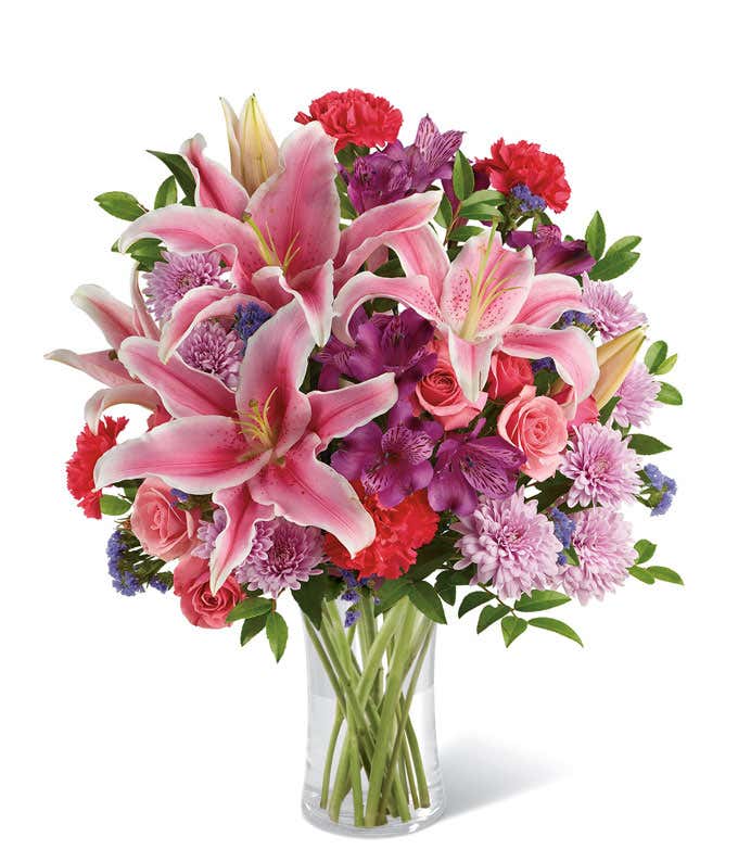 A vibrant bouquet of fresh pink lilies, red carnations, lavender chrysanthemums, purple alstroemeria, pink roses, and greenery arranged in a clear glass vase. The lush mix of colors creates a cheerful and elegant floral display.