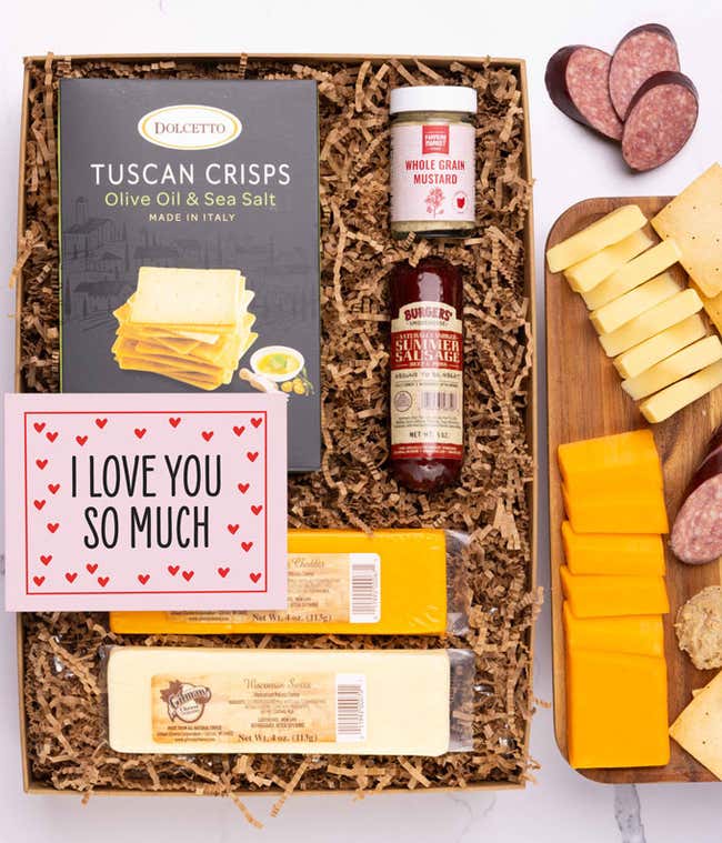 A gift box filled with two different hard cheeses, a small jar of stone-ground mustard, gourmet crackers, a summer sausage, and a pink card with hearts that reads 'I Love You So Much'
