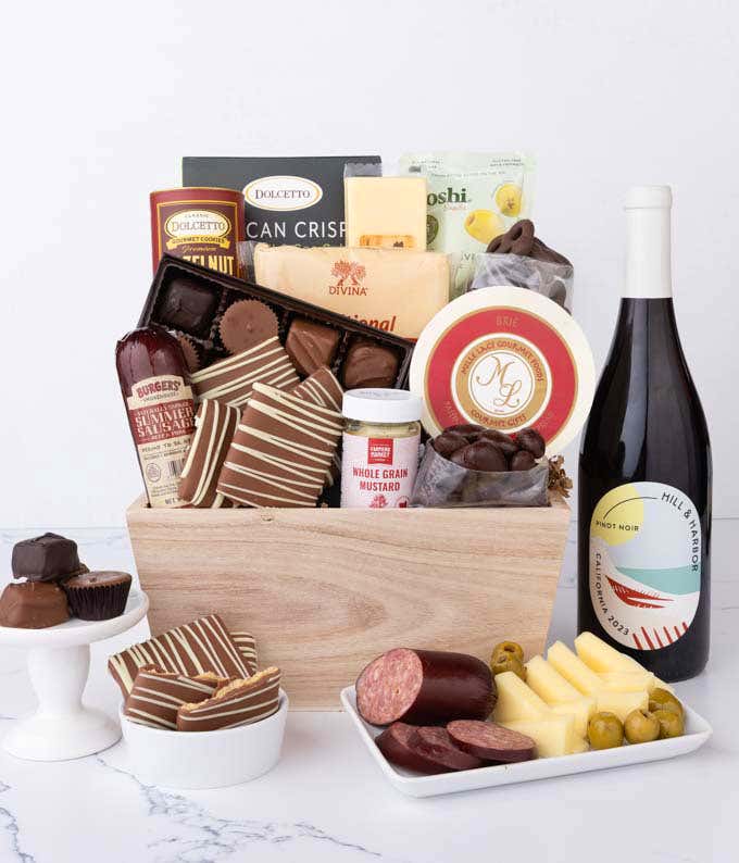 Wooden crate filled with gourmet snacks and a bottle of red wine, including summer sausage, creamy cheeses, Sparkling Winechocolate-covered grahams and almonds, assorted chocolates, wafers, crisps, mustard, and olives, styled with plates of cheese and sau