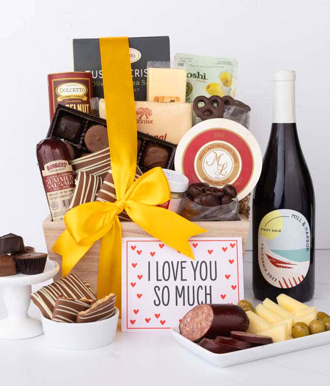 A wooden basket filled with chocolates, cheese, sausage, crackers, crisps, and snacks, paired with a bottle of red wine and an I Love You card, with chocolates and charcuterie displayed.
