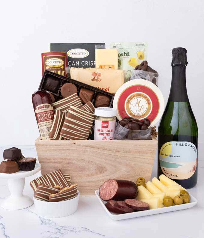 Wooden crate filled with gourmet snacks and a bottle of sparkling wine, including summer sausage, creamy cheeses, chocolate-covered grahams and almonds, assorted chocolates, wafers, crisps, mustard, and olives, styled with plates of cheese and sausage.