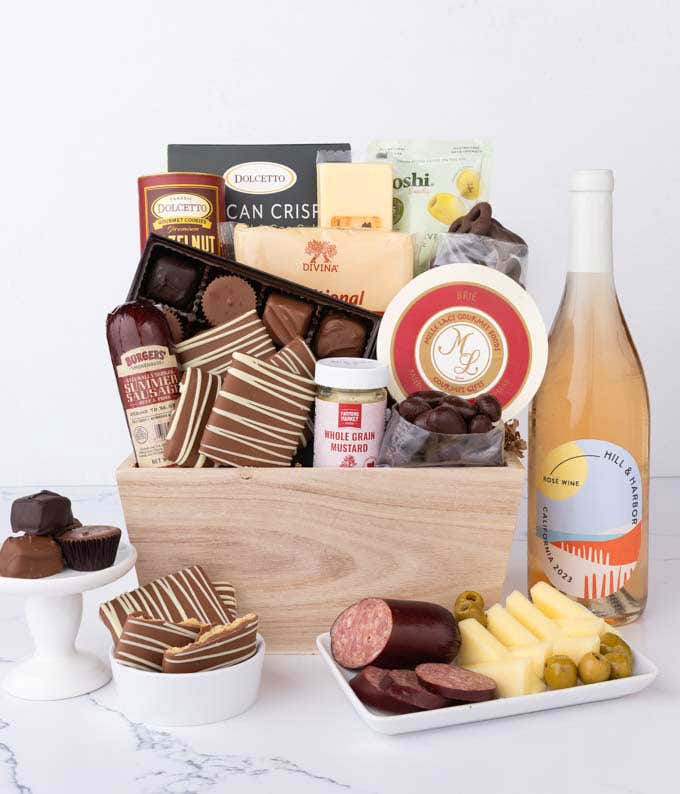 Wooden crate filled with gourmet snacks and a bottle of rosé wine, including summer sausage, creamy cheeses, chocolate-covered grahams and almonds, assorted chocolates, wafers, crisps, mustard, and olives, styled with plates of cheese and sausage.