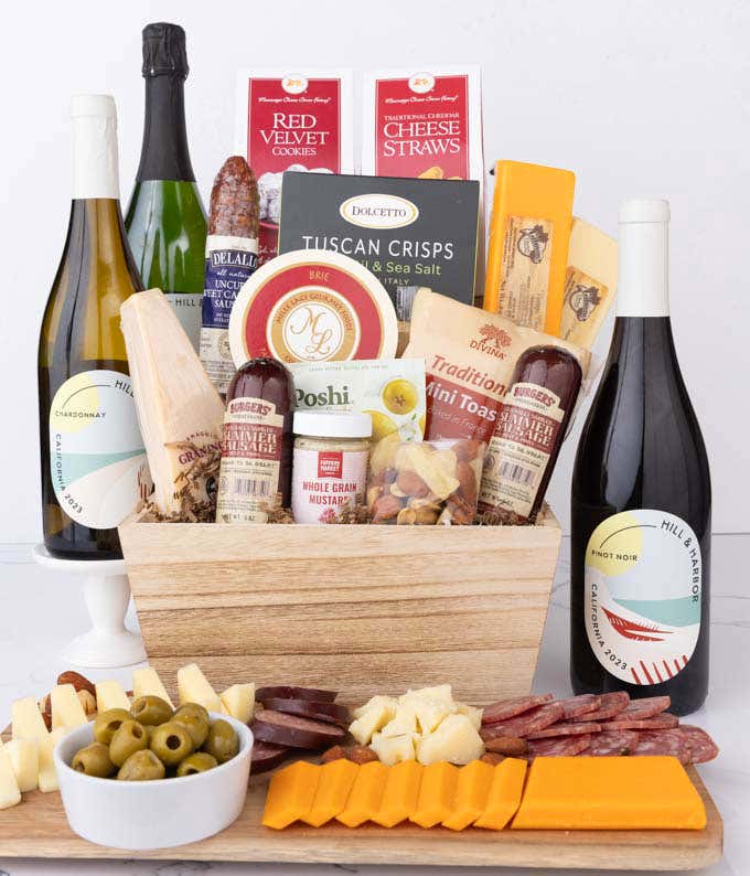 Wooden basket filled with 4 cheeses, 3 sausages, crackers, cookies, nuts, mustard, olives and crisps alongside 3 bottles of wine - Pinot Noir, Chardonnay & Sparkling Wine.