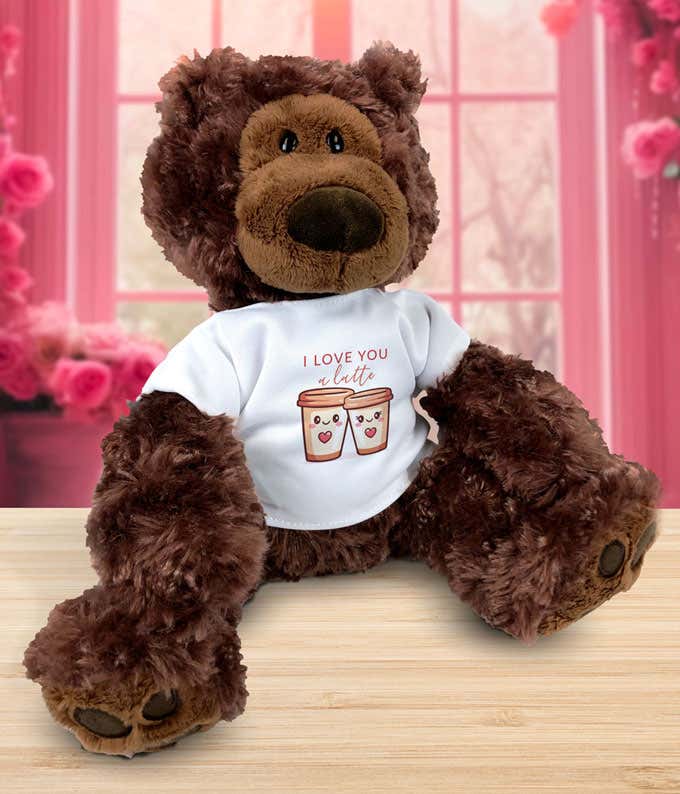 Plush brown teddy bear sitting on a wooden surface, wearing a white T-shirt that reads ‘I love you a latte’ with an illustration of two smiling coffee cups, set against a pink, rose-filled background.