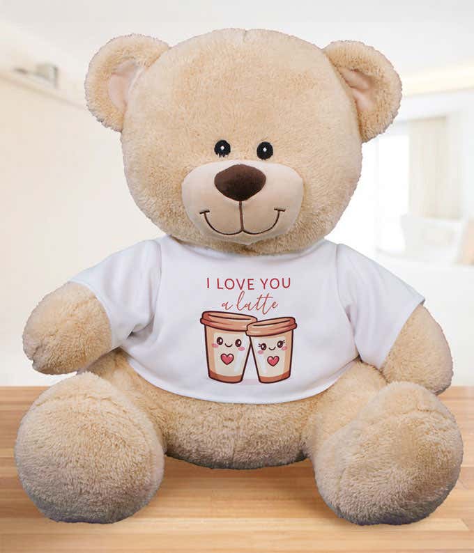 Light tan plush teddy bear sitting on a wooden surface, wearing a white T-shirt that reads ‘I love you a latte’ with an illustration of two smiling coffee cups.